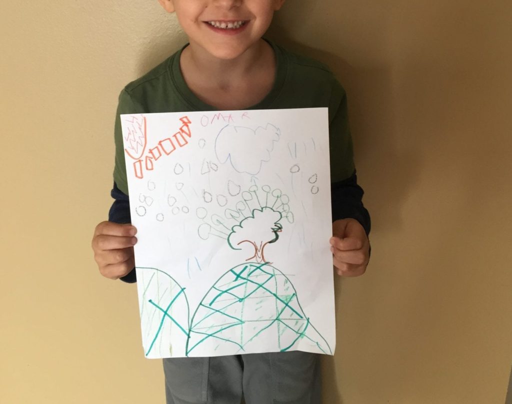 A student holding a drawing of a landscape with patterns drawn in the hill, tree, sky, and sun