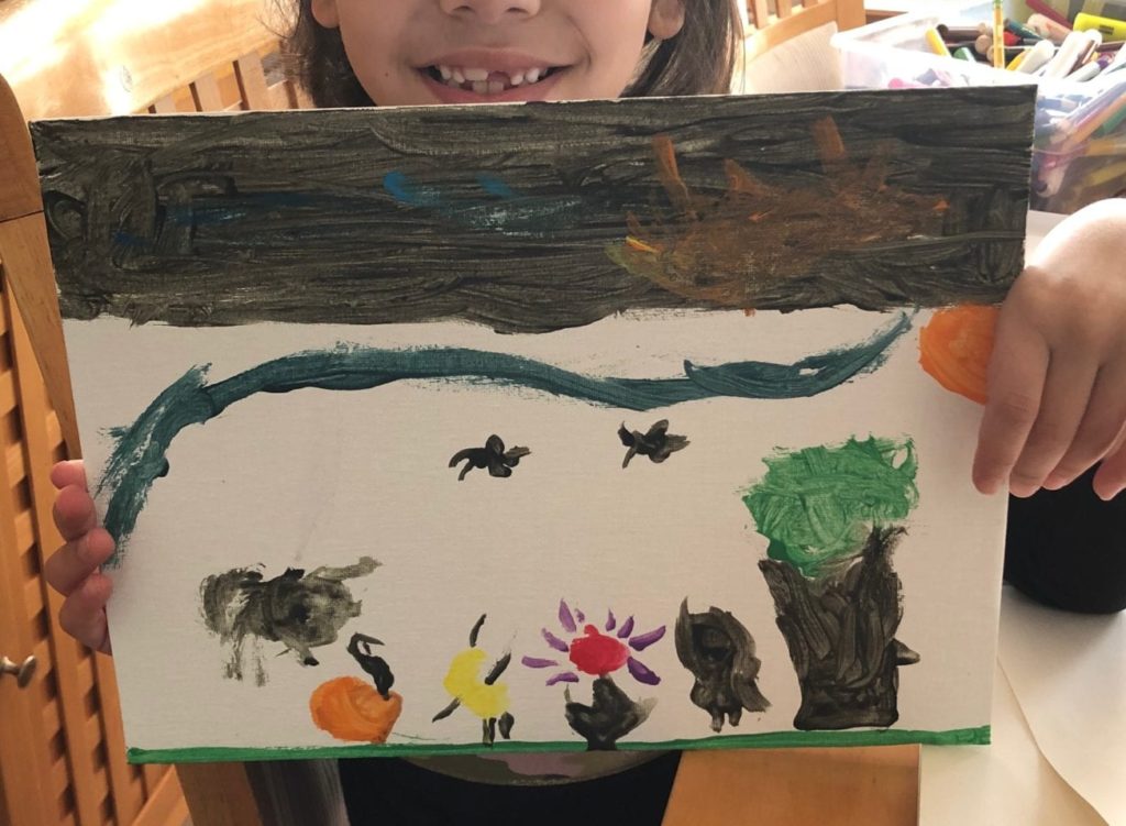 A students is holding a painting sa made; it shows a dark sky on top, then the sky turns white and a tree, flowers, and animals are shown on the ground.
