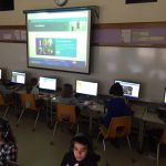Students working on desktop computers. Lesson projected on screen in front of computer lab.