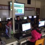 Students working on desktop computers. Lesson projected on screen in front of computer lab.