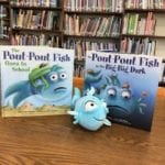 two books and blue stuffed toy fish displayed on a library shelf