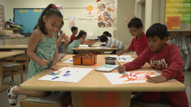 Two boys and a girls students are in the foreground at a wood table painting with watercolor paints. Another table of students are painting in the background.