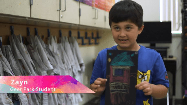 A boy student is showing his drawing of a colorful rocketship on a black background.