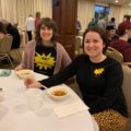 Two woman with 'art is my superpower' shirts on are eating soup.