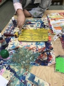 Image of student's hands drawing lines with a fork into wet paint with different materials on the table around them