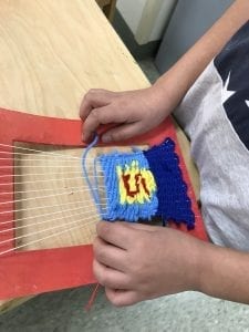 student hands weaving a tapestry tat looks like the super man logo with a yellow pentagon with a red "S" in the middle and blue all around it