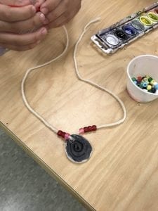 student hands working on threading beads onto a necklace; the necklace is symmetrical and has a ceramic coil in the middle and pink and red beads on either side of the coil