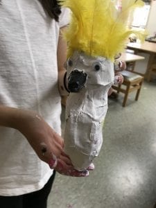 student holding her bird; it is painted white, had 2 googly eyes, a black beak, and yellow feathers on top of its head