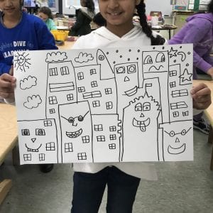 Students drew their buildings in Sharpie marker first. Here is a student holding up her artwork with several buildings drawn in Sharpie; each building has a unique face and windows