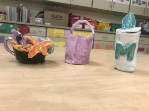 three student made ceramic pieces; one looks like a bowl with flowers around the rim, the other is a purple cylinder with a purple handle over the top and the third is a white cylinder with a teal "M" and a lid with a teal leaf
