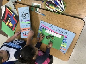 two students working at a table; one is coloring their green folder and the other is putting his artwork into his folder