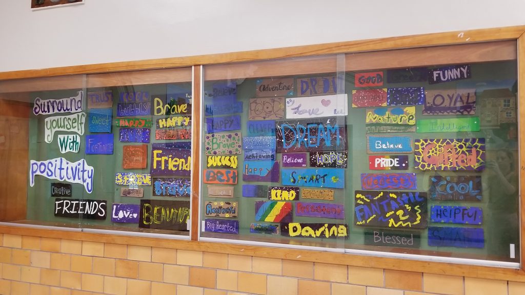Glass display case filled with colorful pieces of cardboard, of all sizes and shapes, with a variety of positive words painted on them.