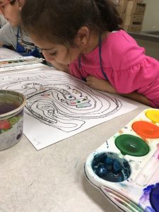 Three students are sitting at a table with white paper painting the lines and shapes they drew while listening to jazz.
