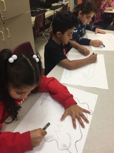 Three students are sitting at a table with black crayons and white paper drawing lines and shapes.