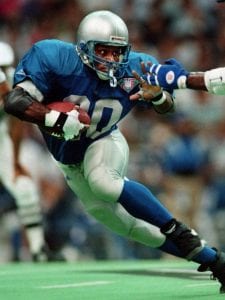 image of Sanders wearing a Lions football jersey holding a football, running, and leaning away as an arm is stretched out towards him from another player