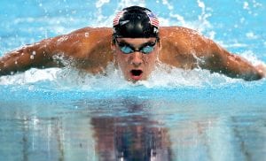 Phelps swimming in the water; he has a swim cap and goggles and the top of his torso and arms and head are out of the water as he takes a breath