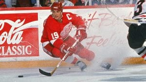 Yzerman skating wearing a Red Wings jersey, he is holding his stick out in front of him going forward a puck