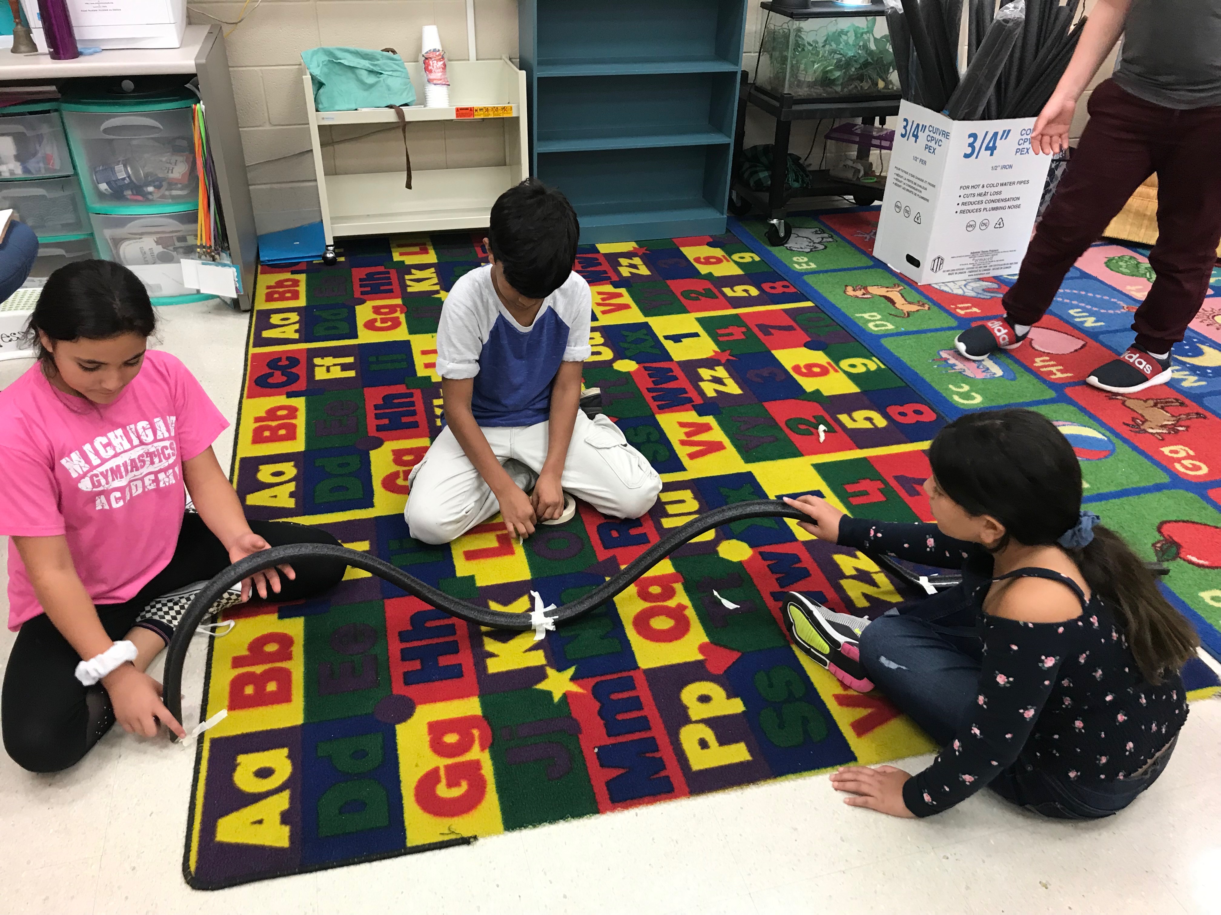 Students sitting on carpet, building a model roller coaster.