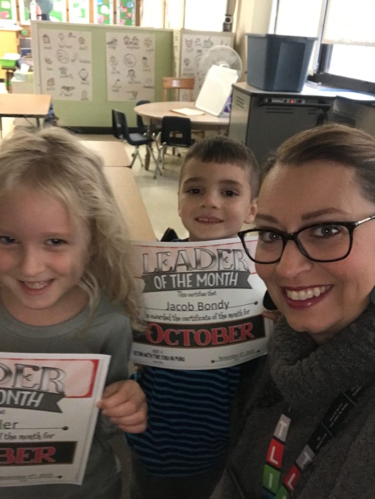 two students smile with their teacher while holding their leader of the month certificates