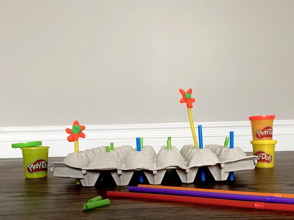 Picture of completed activity. Straws sticking out from egg carton with playdoh flowers. Materials surrounding image.