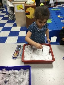 A child using a paint brush in a tray of snow, with a set of watercolor paint next to the tray.
