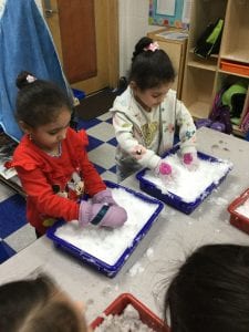 Two children standing at a table. Each wearing gloves and putting their hands into a tray of snow.