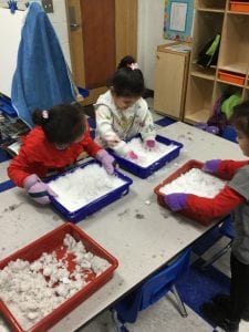Three children standing at a table, each with their own tray of snow. Two are holding the side of the trays, while the other is putting her hands into the snow.