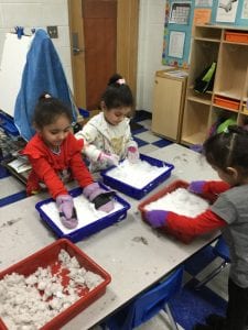 Three girls standing a table, each with their own tray of snow. Two girls have their hands in the tray of snow, the other is holding the sides of the tray.